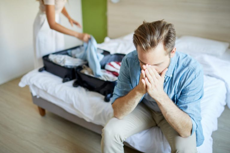Young man sitting on bed and praying while his wife getting suitcase before leaving