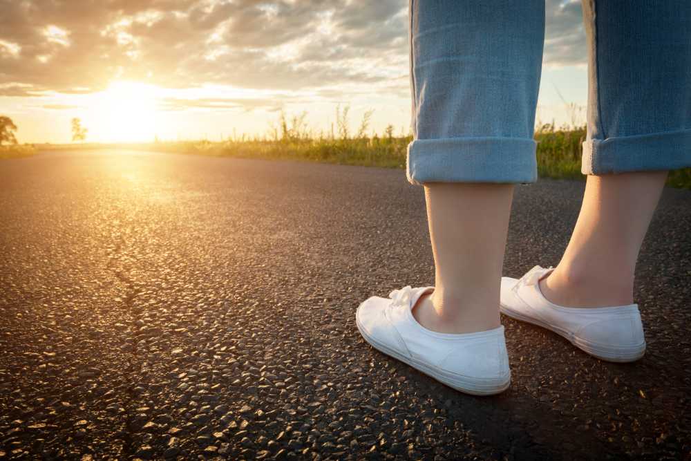 Woman in white sneakers standing on asphalt road towards sun. Concept of new start, travel, freedom etc.