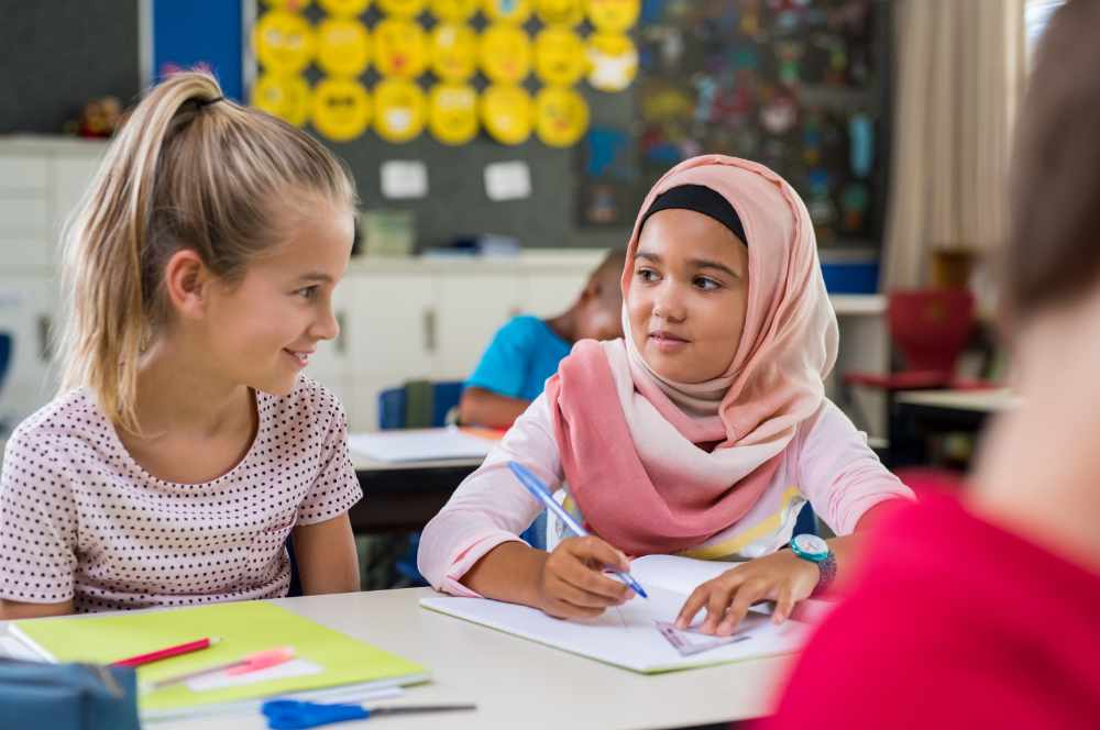 Young arab girl with hijab doing exercise with her bestfriend at international school. Asian muslim school girl sitting near her classmate during lesson. Multiethnic elementary students in classroom.