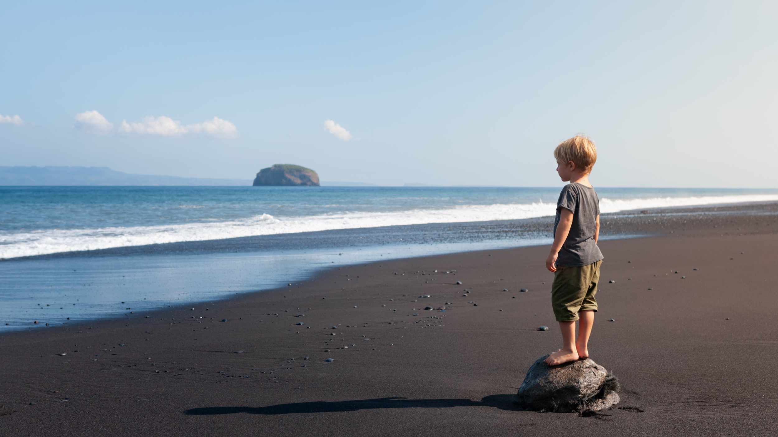 Little kid stand on big stone on black sand sea beach. Dreaming child look at sea surf, waves. Solitude concept. Retreat leisure on summer family vacation