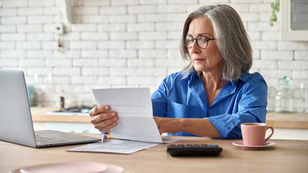 Adult senior 60s woman working at home at laptop. Serious middle aged woman at table holding document calculating bank loan payments, taxes, fees, retirement finances online with computer technologies