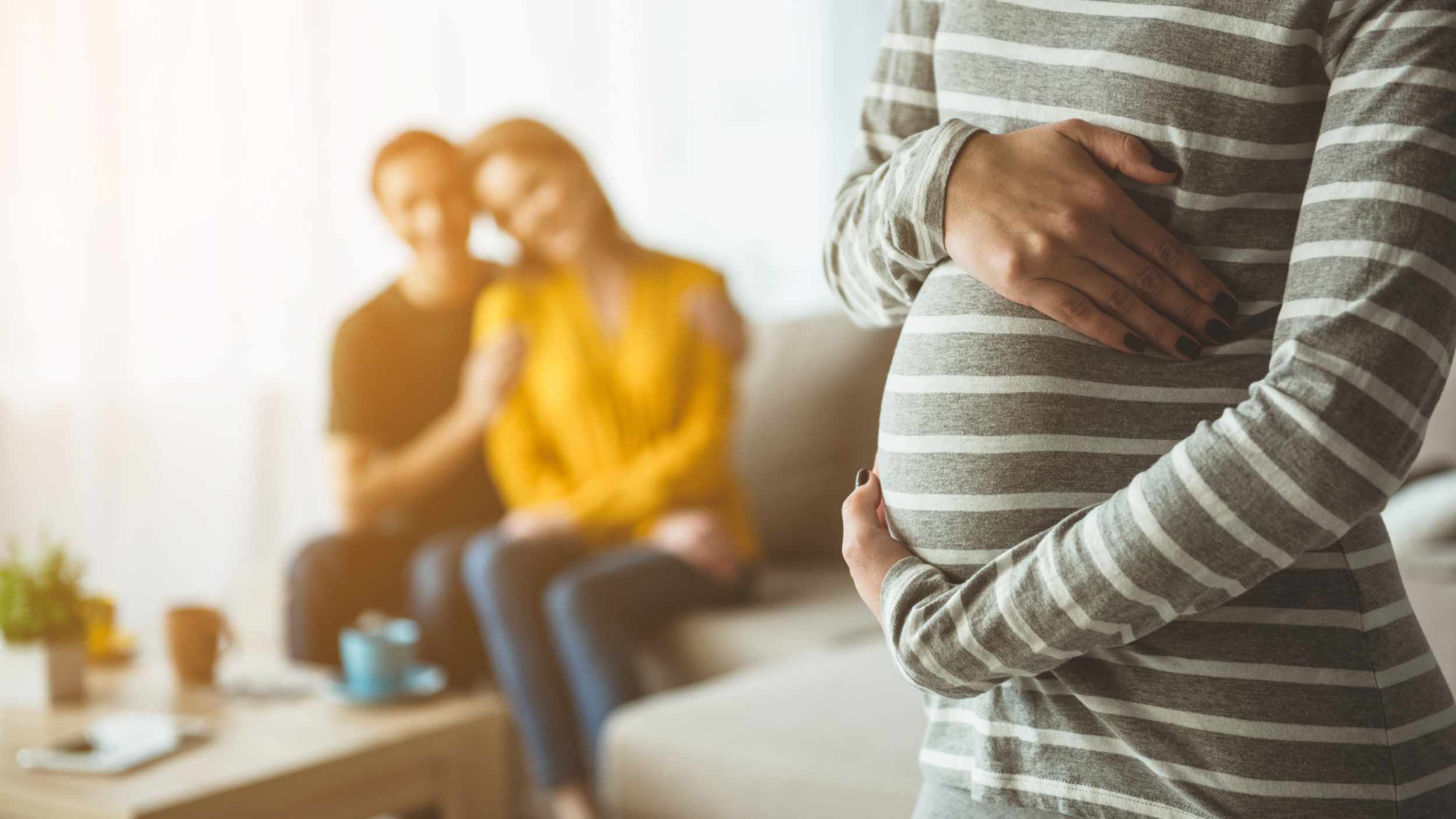 Focus on pregnant woman touching her belly. Happy married couple are sitting on sofa and embracing on background. Surrogacy concept