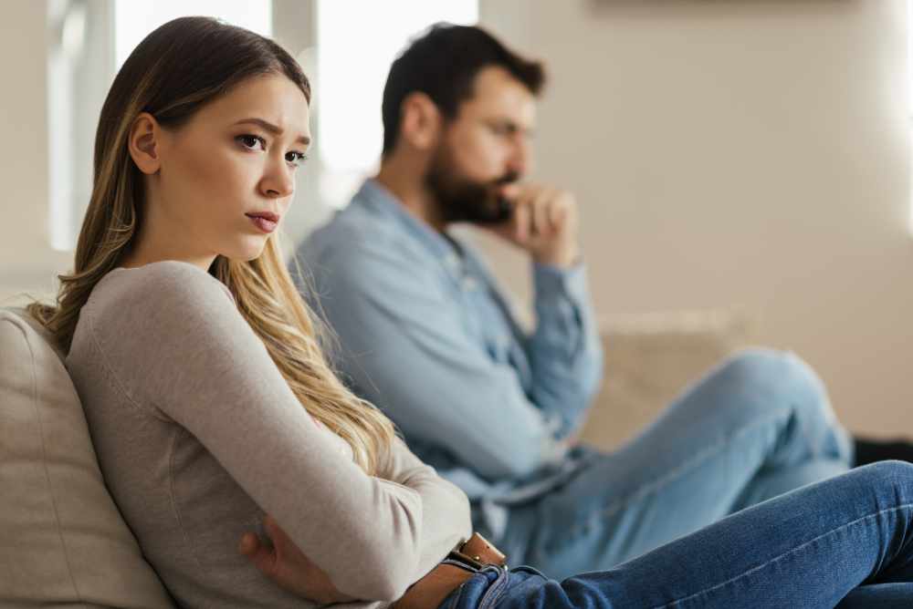Worried young woman sitting on sofa at home and ignoring her partner who is sitting next to her