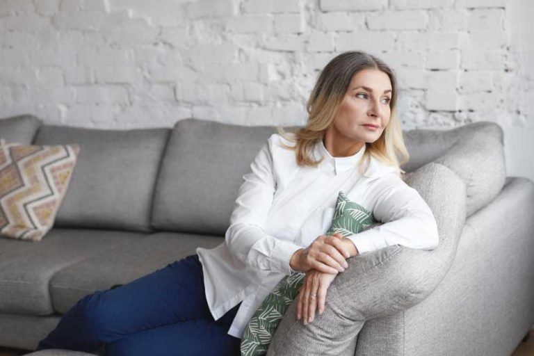 People, rest, leisure and relaxation concept. Candid shot of gorgeous elegant sixty year old mature female with loose gray hair relaxing at home, having pensive look, sitting comfortably on sofa